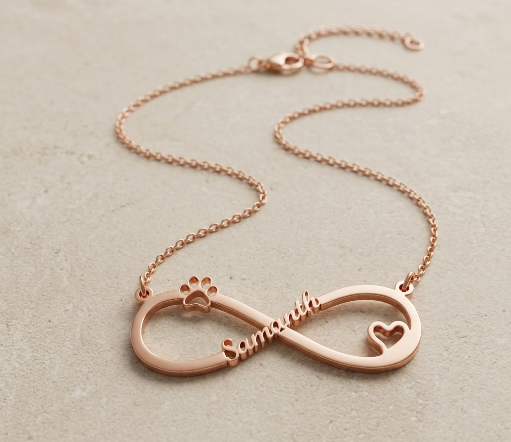 A clean flat lay product photo of a rose gold infinity pendant necklace on a smooth warm-toned grey concrete or stone surface. The infinity-shaped pendant is engraved with the name "Samantha" in raised cursive lettering running through the center crossbar. The left loop of the infinity symbol features a raised paw print cutout at the top, and the right loop contains a small open heart cutout. The pendant connects to a fine rose gold cable chain that curves naturally into an S-shape across the surface, endin